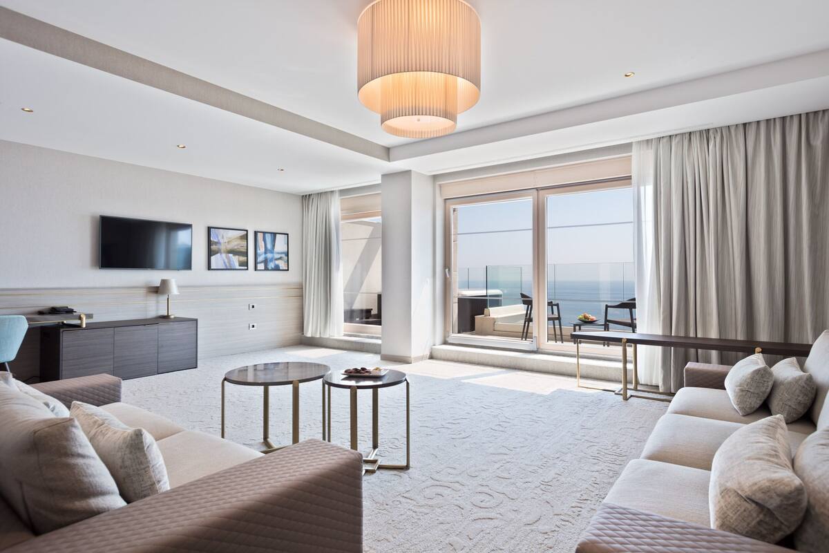 Una habitación de hotel moderna con un balcón grande, un televisor de pantalla plana, un sofá, una mesa de centro y una zona de comedor con vista al océano.