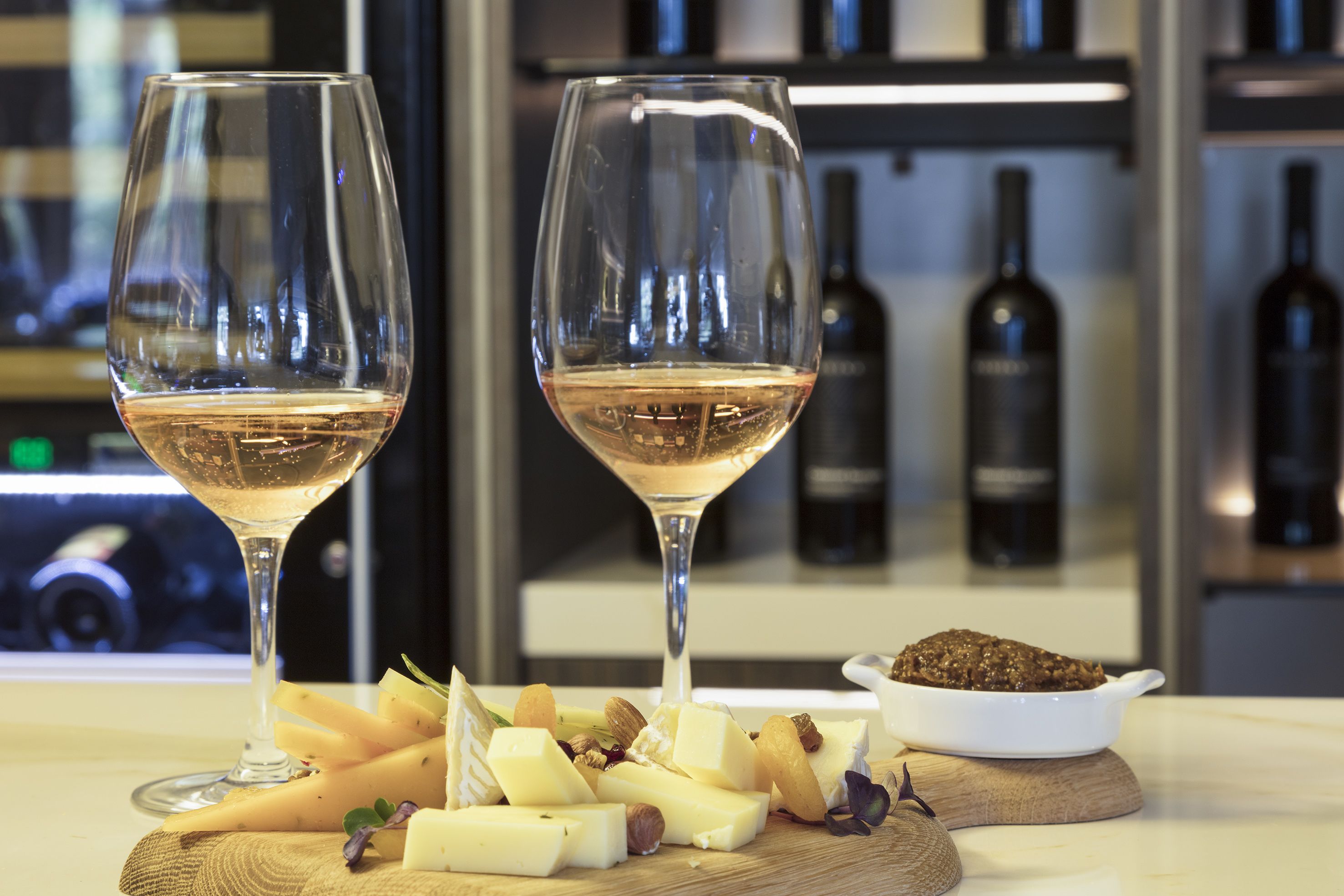 Dois copos de vinho branco, uma tábua de madeira com queijo e frutos secos, e uma tigela de creme de chocolate.