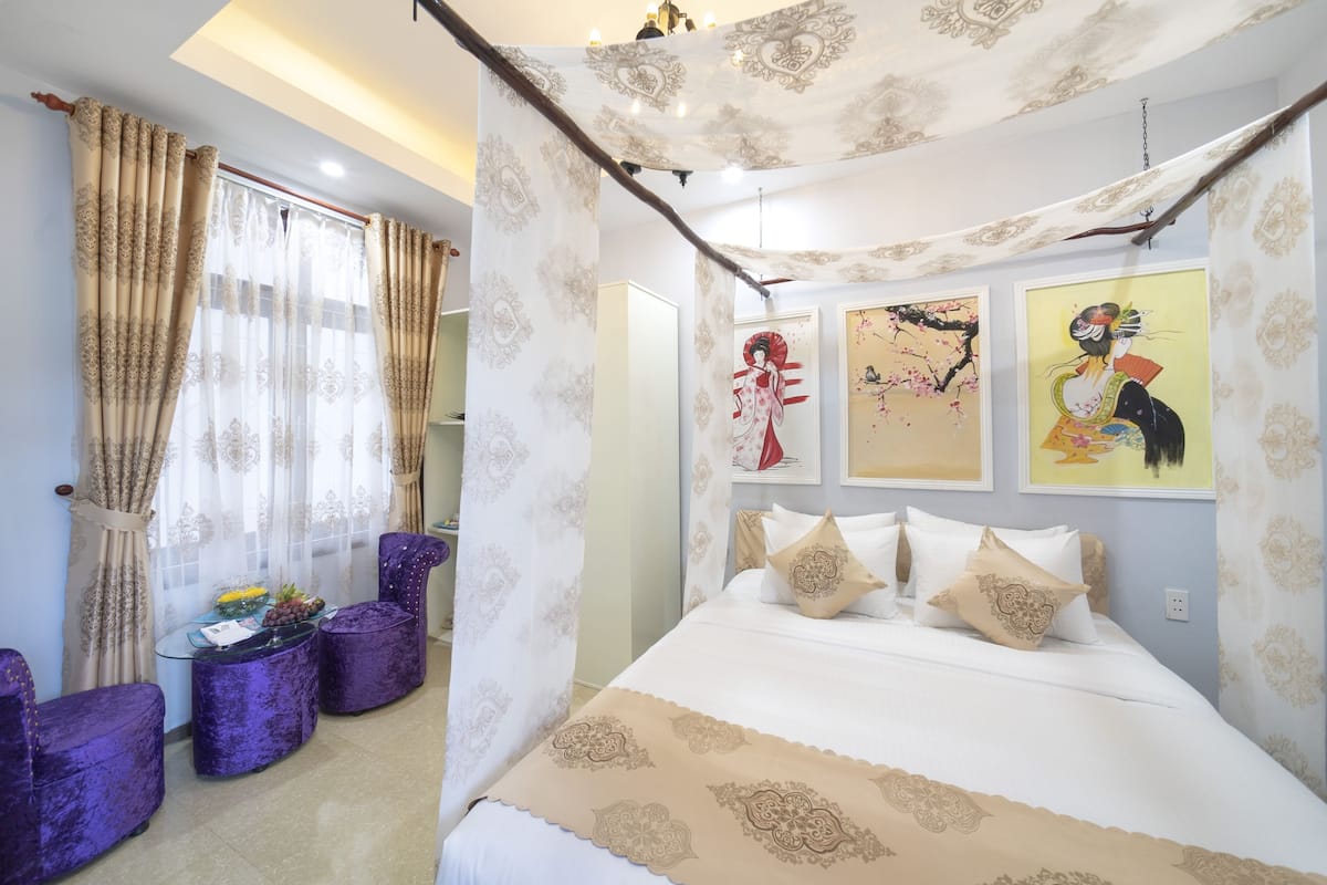 A bedroom with a bed, a table with fruits, purple chairs, and artwork on the wall.