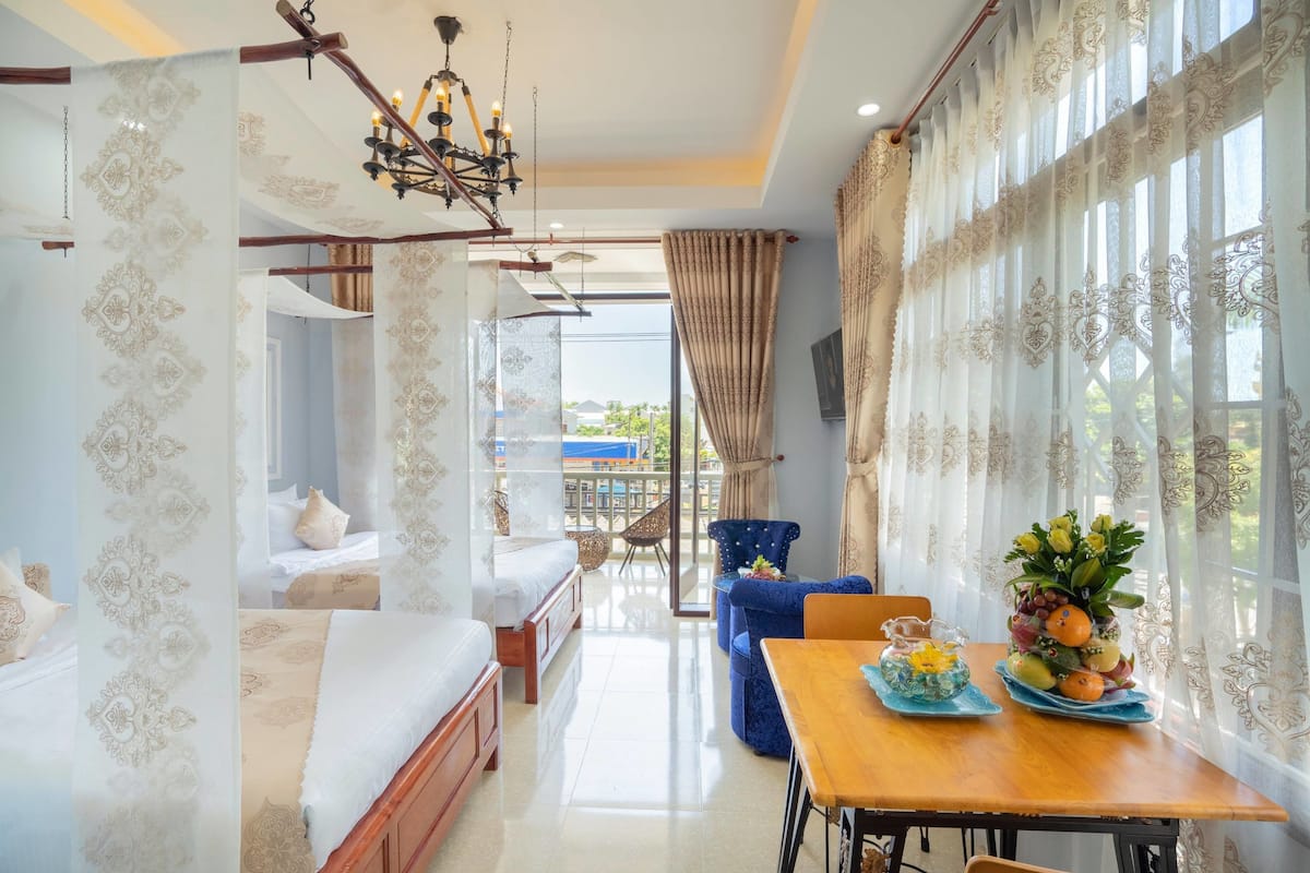 A hotel room with a bed, a dining table with fruit, a chandelier, and a balcony with a view.