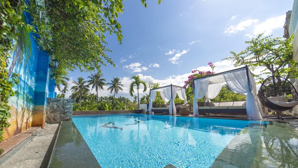 A clear swimming pool with a stone wall on one side, surrounded by lush greenery and a clear blue sky. - VIP Garden Villa and Pool (Da Nang)