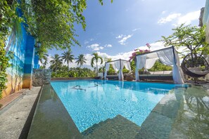 Una piscina cristalina con un muro de piedra en un lado, rodeada de vegetación exuberante y un cielo azul despejado.