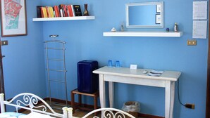 A room with a bed, a desk with a mirror, two shelves with books and decorative items, a suitcase, and a trash bin. - B&B Verdegiada (Brindisi)
