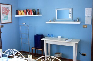 Una habitación con una cama, un escritorio con espejo, dos estantes con libros y objetos decorativos, una maleta y un cesto de basura.