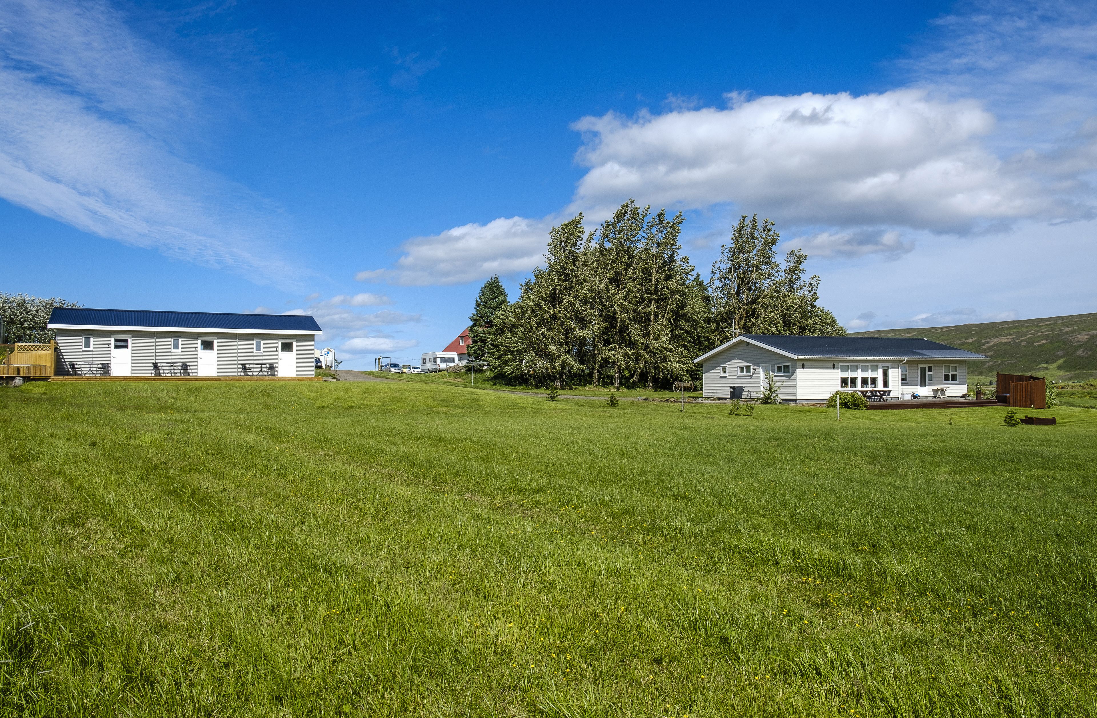 A rural setting with a large grassy field, a white house with a dark roof, and a red-roofed building in the background.
