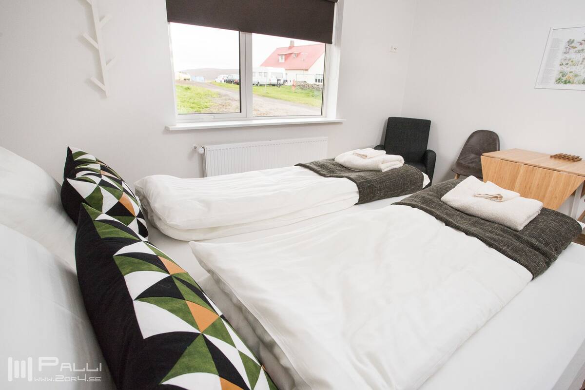 Une chambre d’hôtel avec deux lits, une fenêtre donnant sur une maison, une table de chevet en bois et un radiateur fixé au mur.