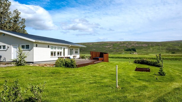 Een wit huis met een terras en een houten constructie in een grasveld, met heuvels op de achtergrond.