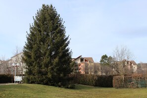 A residential area with a large evergreen tree, a hedge, and a building with multiple balconies. - Hotel Melodie (Bellignat)