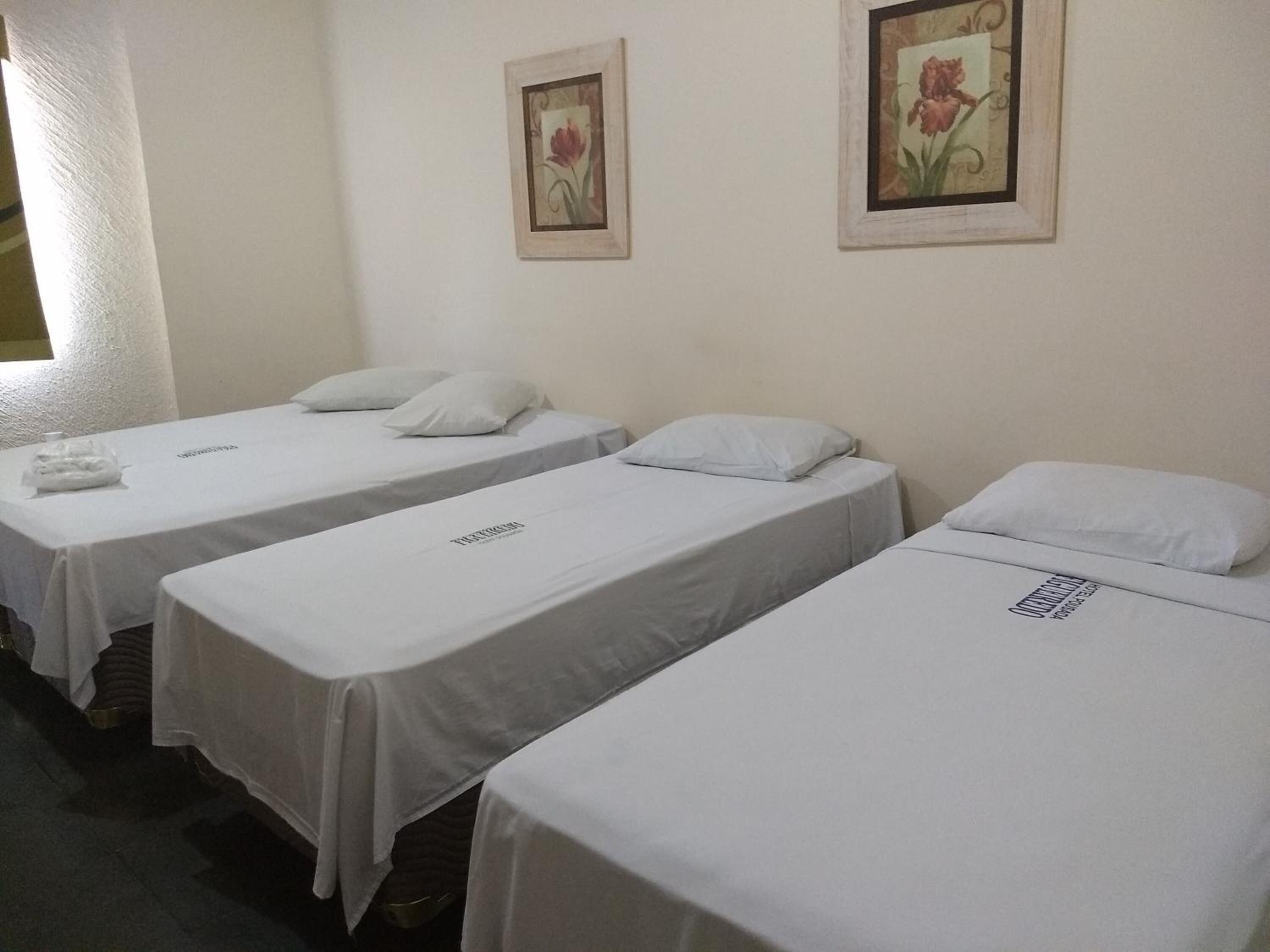 Une chambre d’hôtel avec trois lits simples, chacun recouvert d’une courtepointe blanche et agrémenté d’un oreiller. Des tableaux encadrés représentant des fleurs ornent le mur.