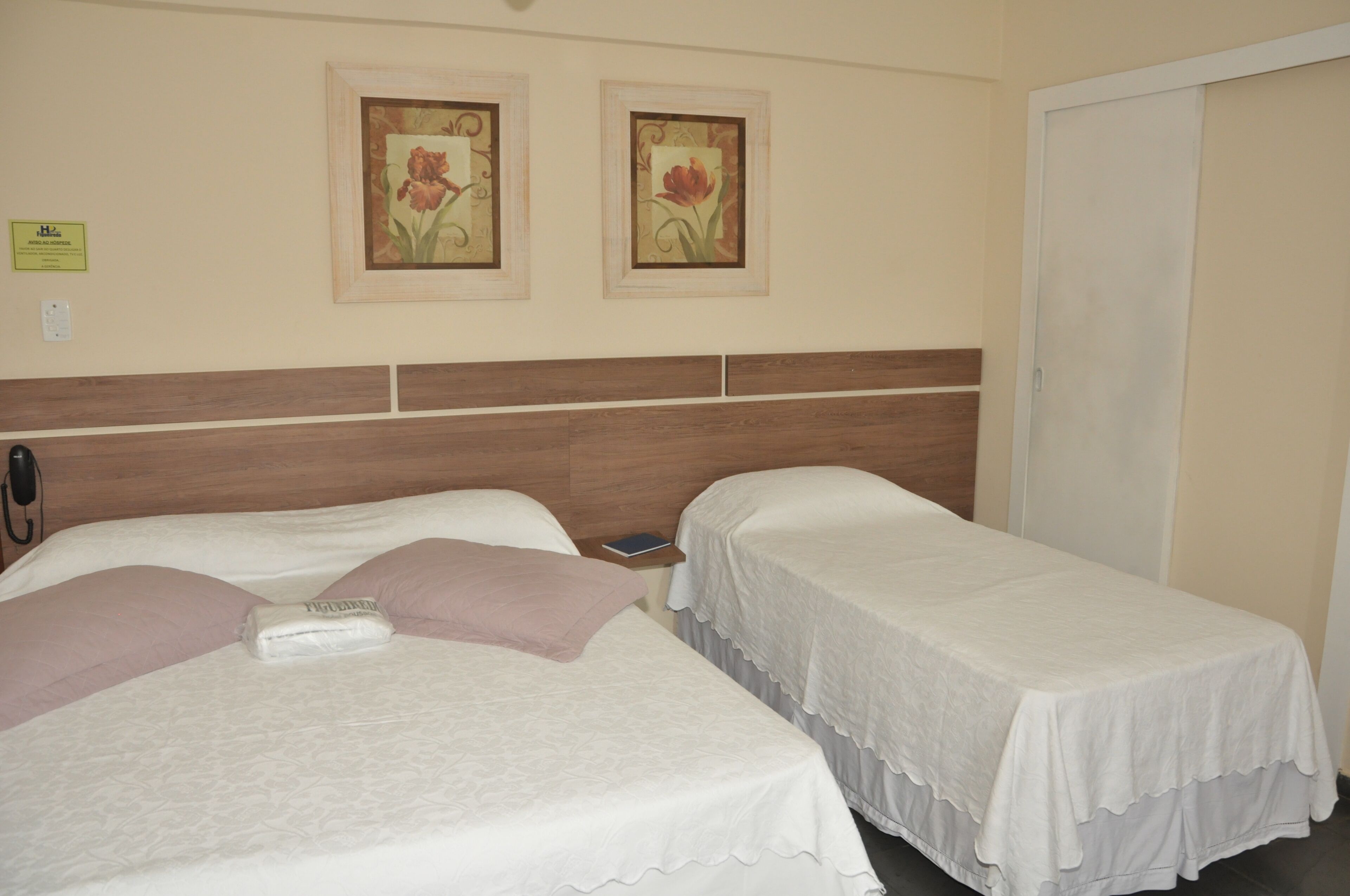 A hotel room with two single beds, a wooden headboard, and two framed floral paintings on the wall.