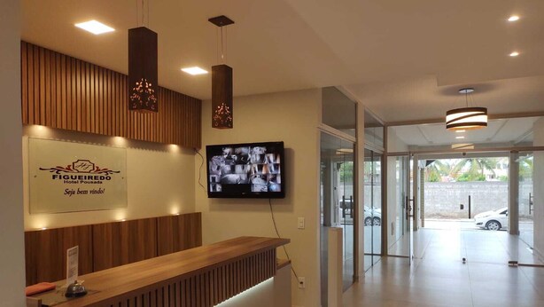 A hotel reception area with a wooden reception desk, a mounted television displaying security footage, and a sign for 'FIGUEIREDO Hotel Pousada'. - Hotel Pousada Figueiredo (Araruama)