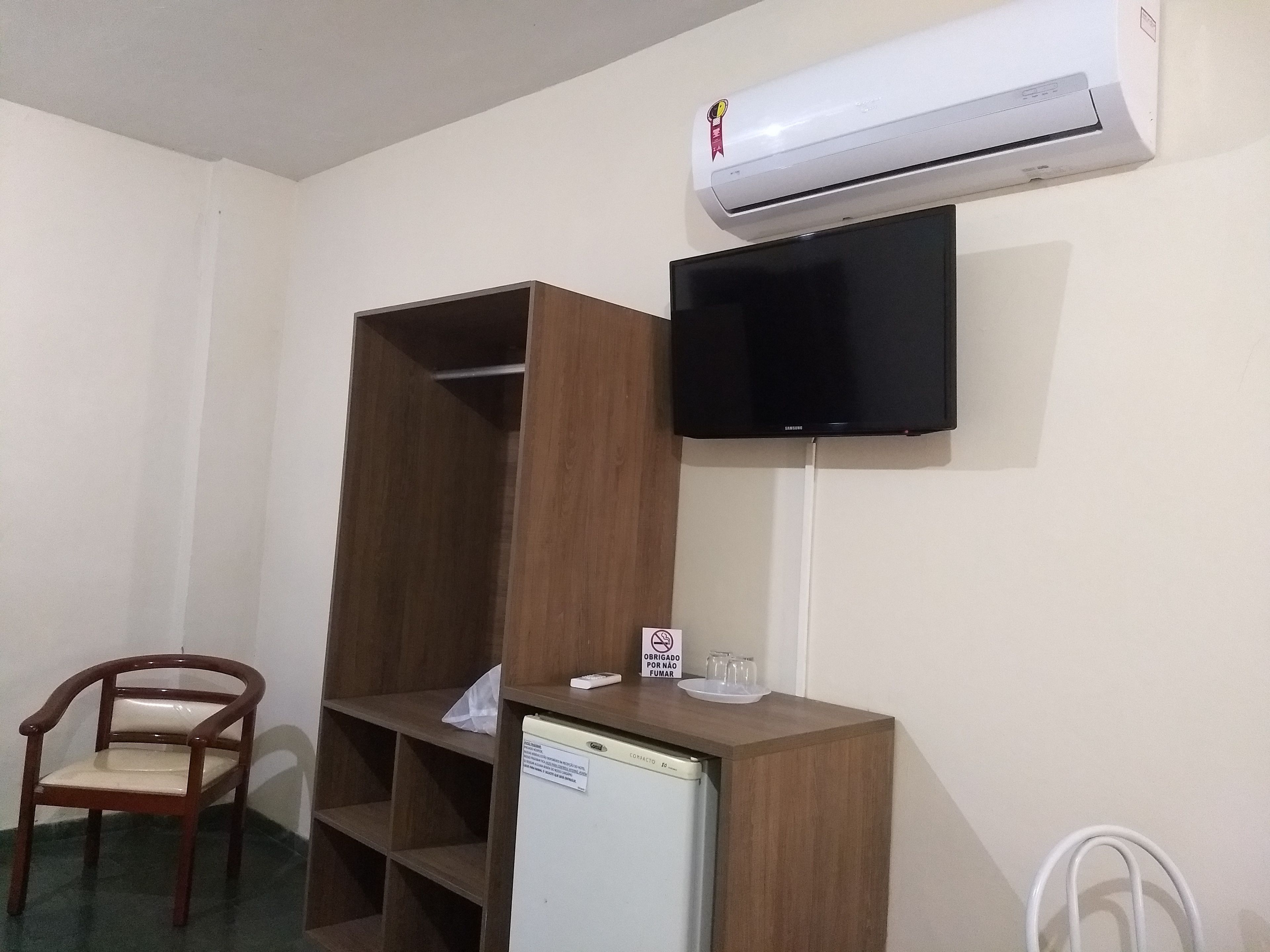 A room with a wooden wardrobe, a television mounted on the wall, a small refrigerator, and a chair.