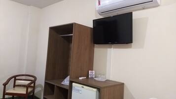 A room with a wooden wardrobe, a television mounted on the wall, a small refrigerator, and a chair.
