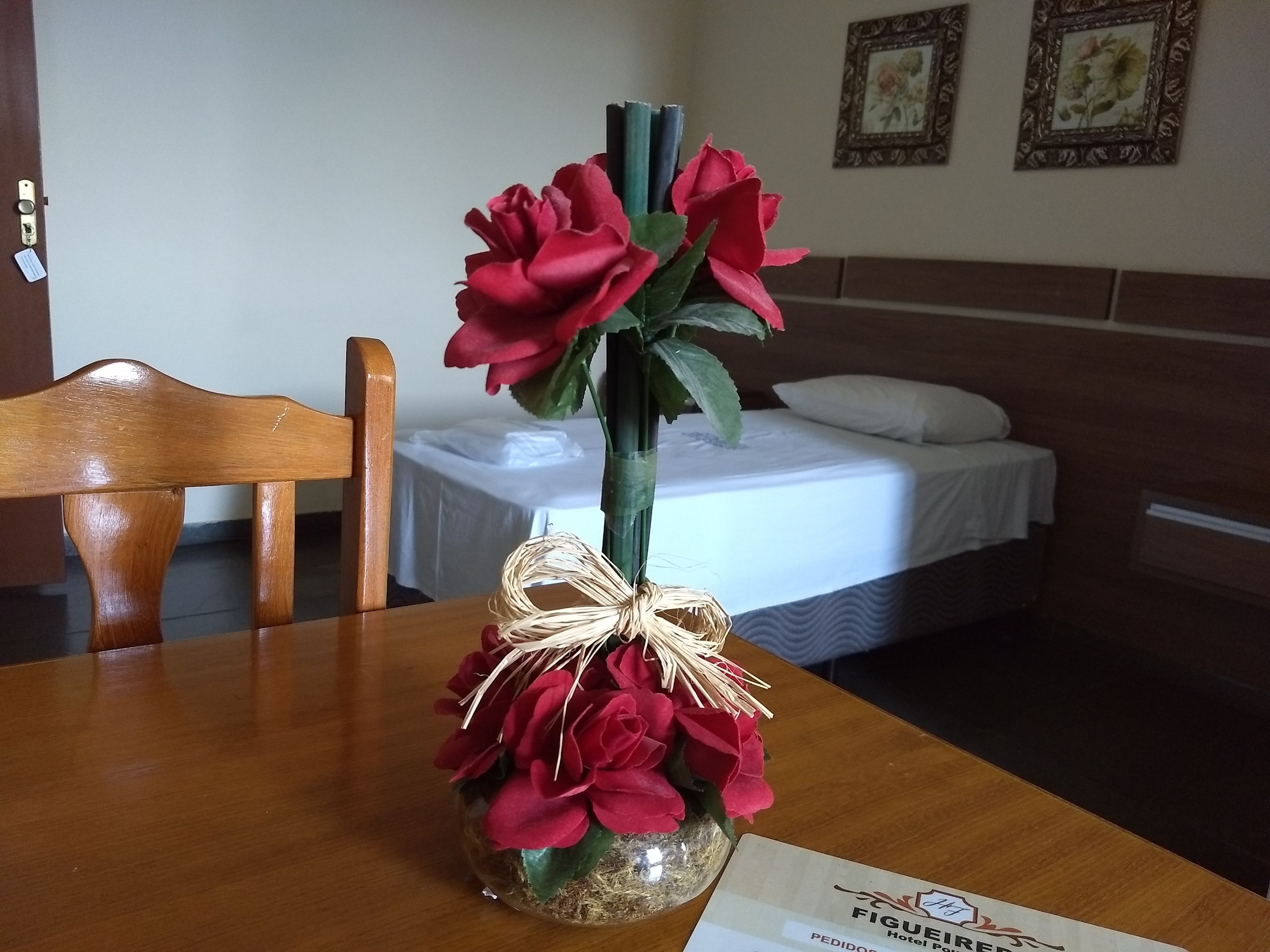 Una habitación de hotel con una mesa de madera, un florero con flores rojas, una cama con ropa blanca y dos cuadros en la pared.