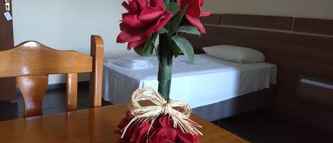 Una habitación de hotel con una mesa de madera, un florero con flores rojas, una cama con ropa blanca y dos cuadros en la pared.
