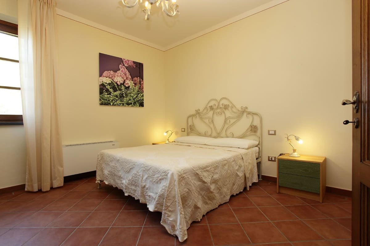Een slaapkamer met een bed, nachtkastje, wandlamp, schilderij en een raam met gordijnen.