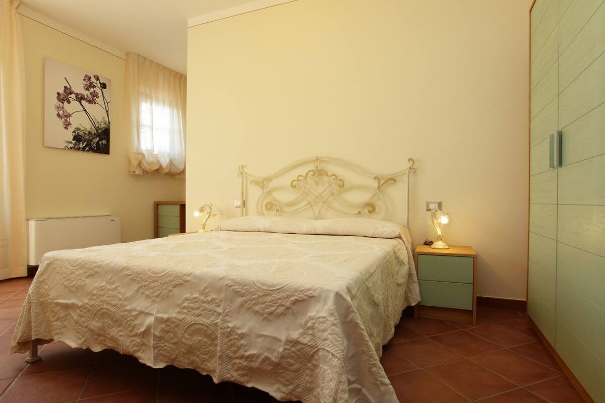 Een slaapkamer met een bed, nachtlampjes, een nachtkastje, een radiator, een aan de muur hangend schilderij en een raam met gordijnen.