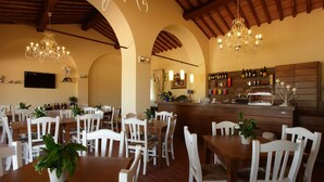 A dining area with wooden tables and white chairs, a bar with bottles, and a TV mounted on the wall. - Agriturismo La Regina (Cascina)