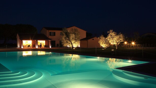 A swimming pool with a lighted edge, a small building with a patio, and trees illuminated by lights. - Agriturismo La Regina (Cascina)