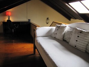 Ein gemütliches Schlafzimmer im Dachgeschoss mit schrägen Wänden, einem Holztisch mit Lampe und einem weißen Sofa mit Streifen-Kissen.