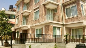 Edifício residencial de vários andares com fachada de tijolos, varandas e um jardim frontal cercado, com grama e vasos de madeira.