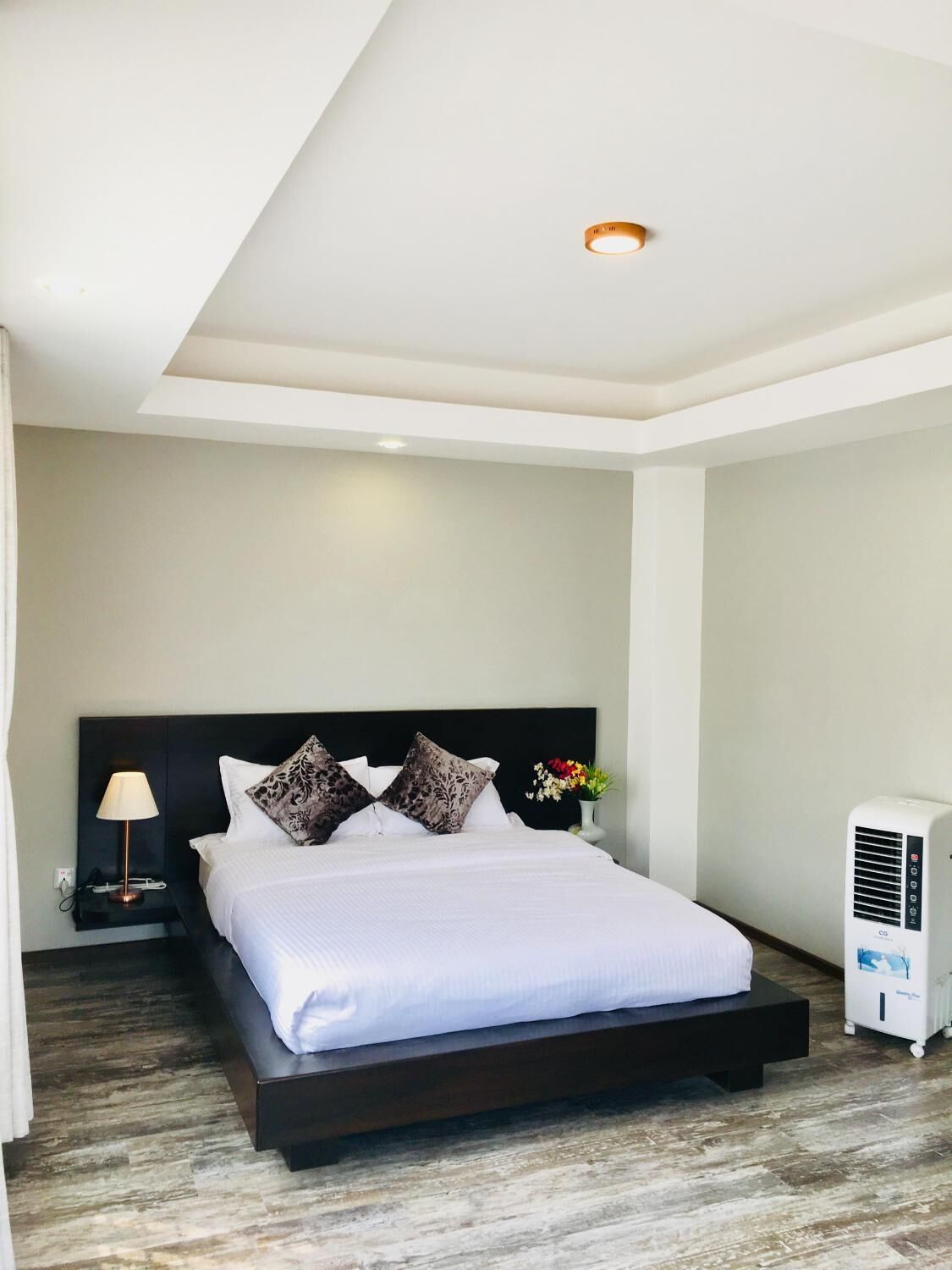 Una habitación de hotel moderna con una cama grande, una mesita de noche con lámpara, otra mesita auxiliar y un aire acondicionado instalado en la pared.