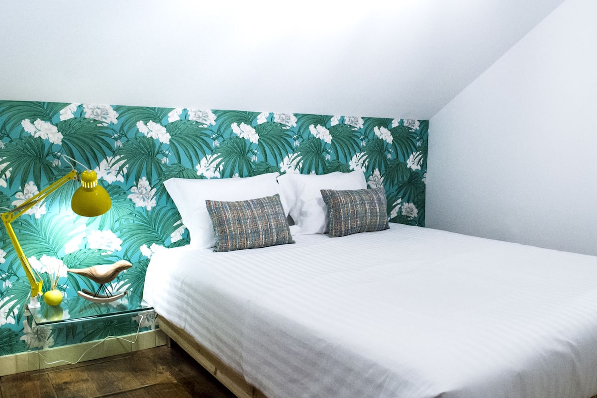 A neatly made bed with white bedding, a glass bedside table with a yellow lamp, and a tropical wallpaper design.