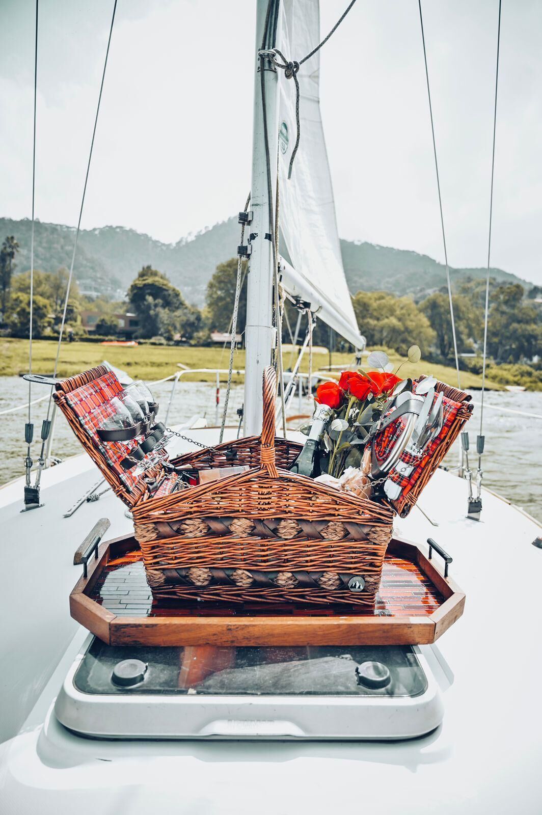 Ein Segelboot mit einem Korbwannenkorb auf dem Deck, einer weißen Segel und ruhigem Wasser im Hintergrund mit Hügeln.
