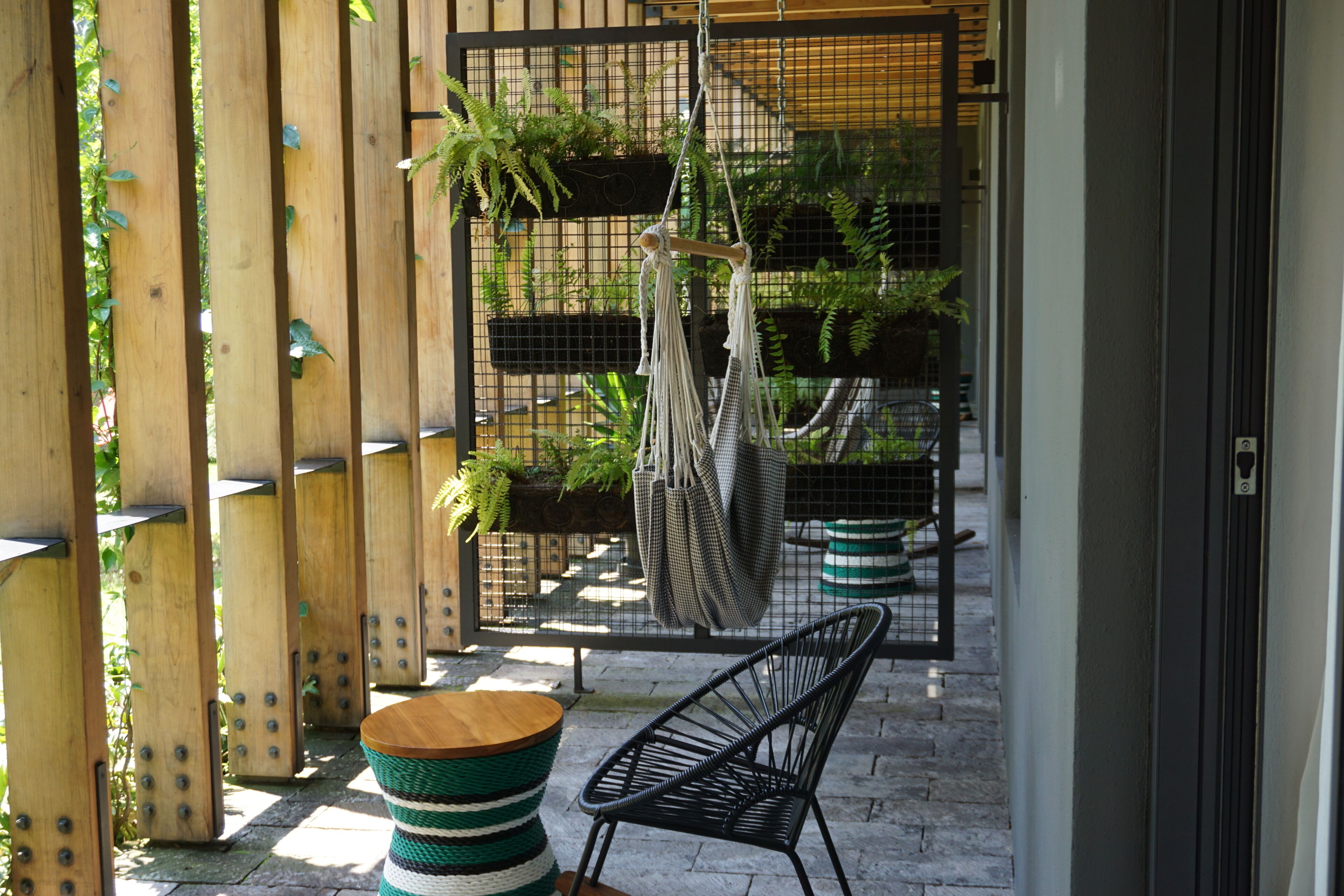 A wooden pergola with hanging macramé chairs and a small round table, surrounded by potted plants and a metal fence.