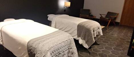 Two beds with white linens, a wall-mounted lamp, a small table with a chair, and a stone-tiled floor.