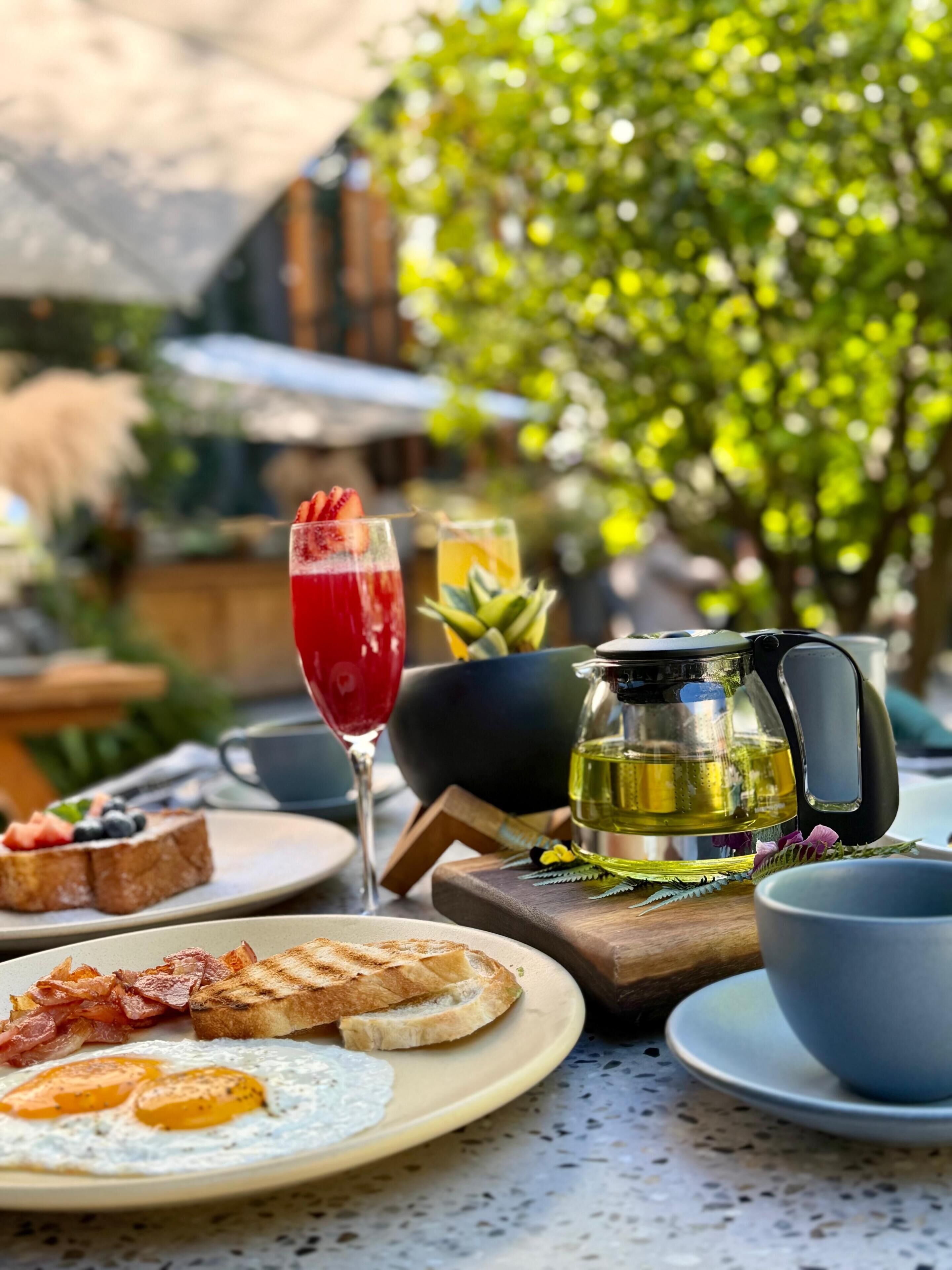 A breakfast setting with toast, eggs, bacon, a red drink, and a pitcher of tea.