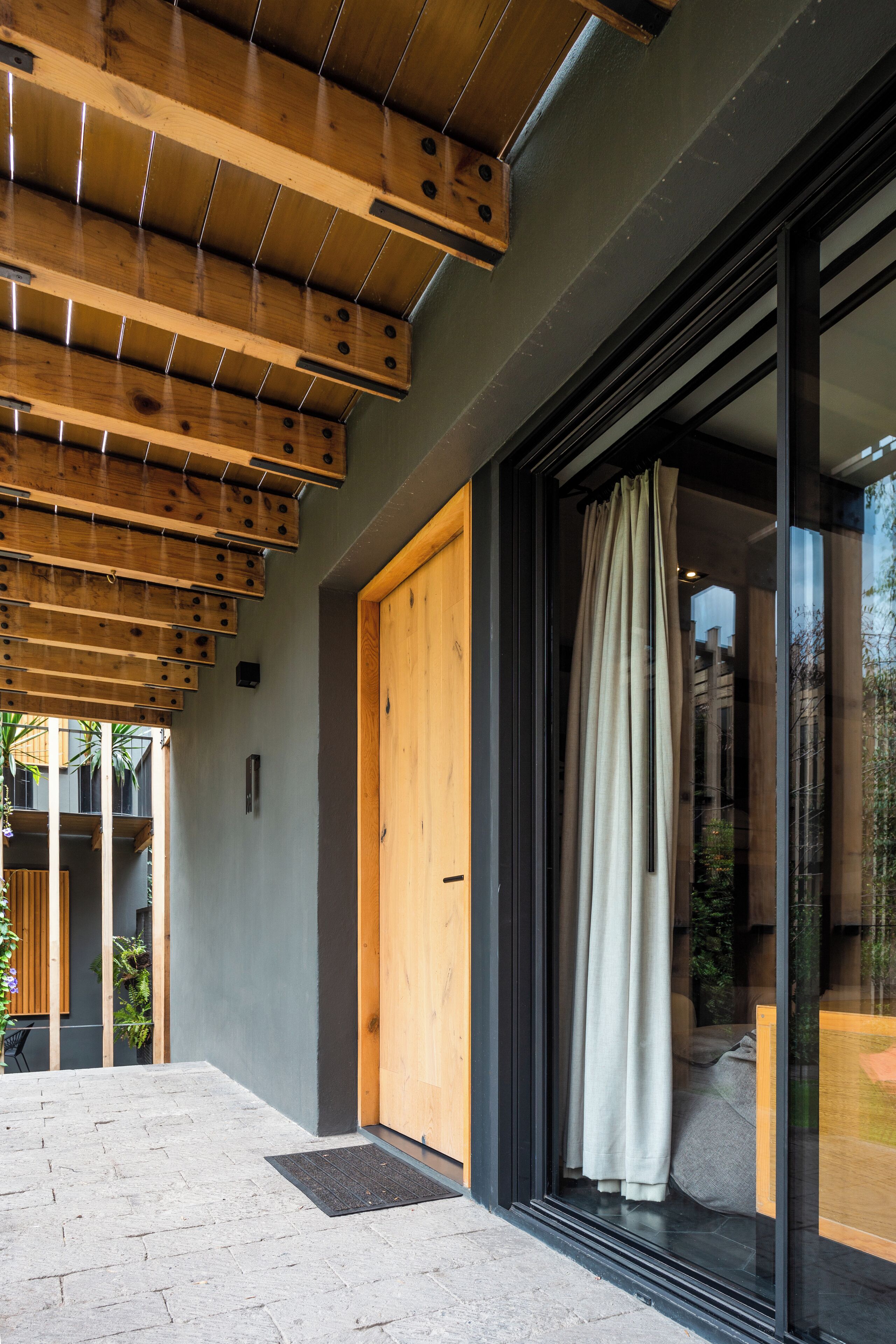 A modern building entrance with a wooden ceiling, a glass door with a curtain, and a paved area with a mat.