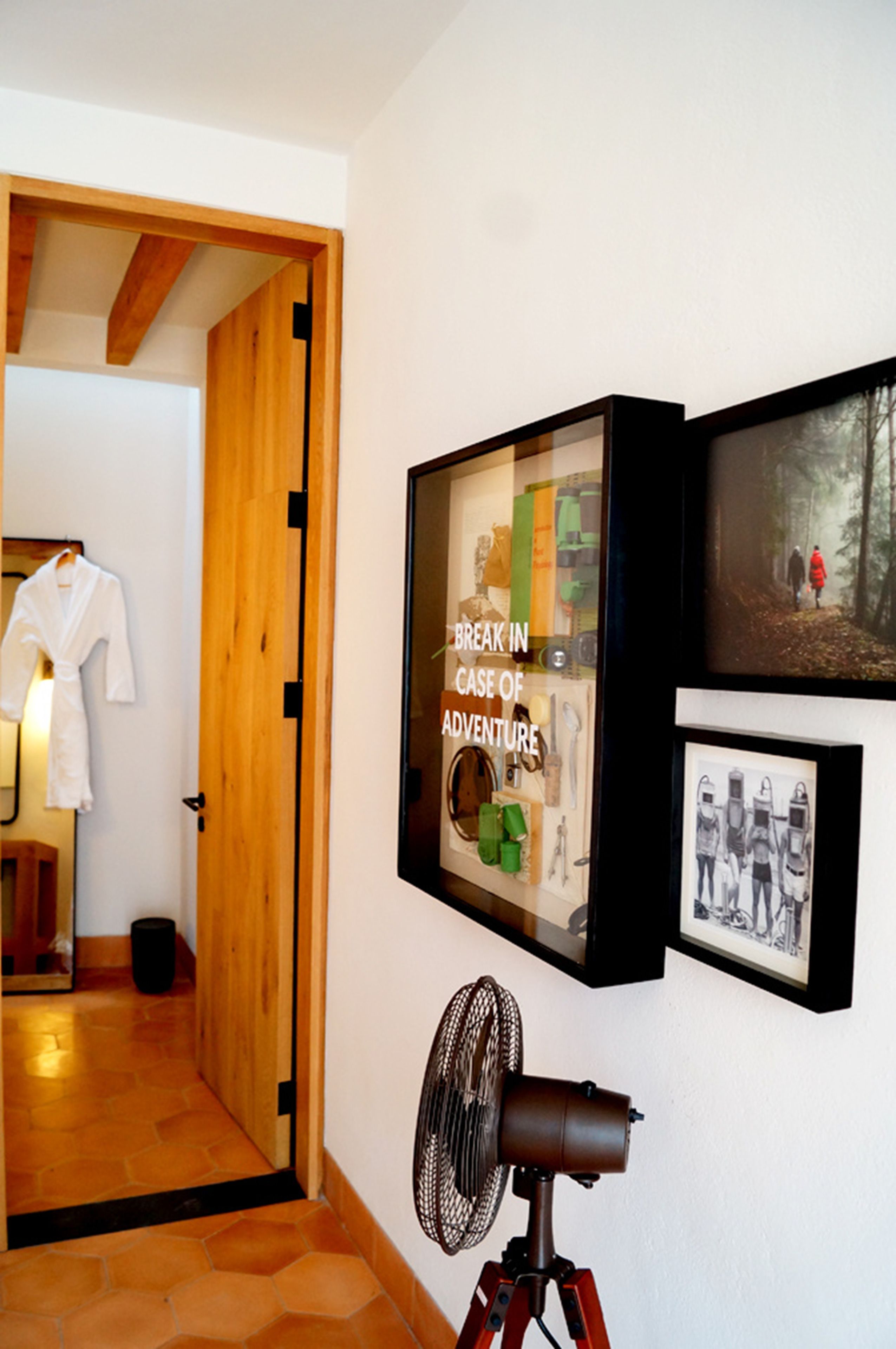 Ein Zimmer mit Holztür, einem Bilderrahmen mit dem Schriftzug "BREAK IN CASE OF ADVENTURE", einem Ventilator und einem gerahmten Foto von Personen.
