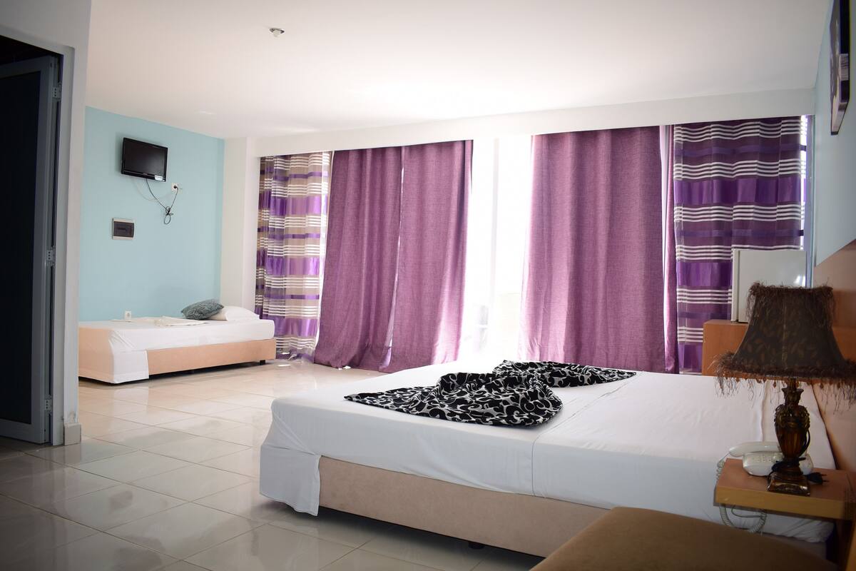 A hotel room with a large bed, a bedside table with a lamp, a TV mounted on the wall, and a window with purple curtains.