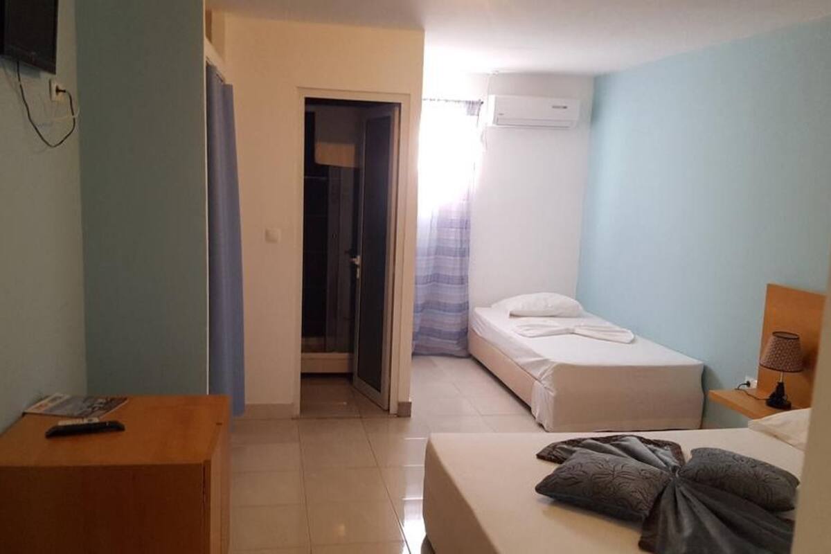 A hotel room with a bed, a desk with a remote control, a door leading to a bathroom, and a window with curtains.