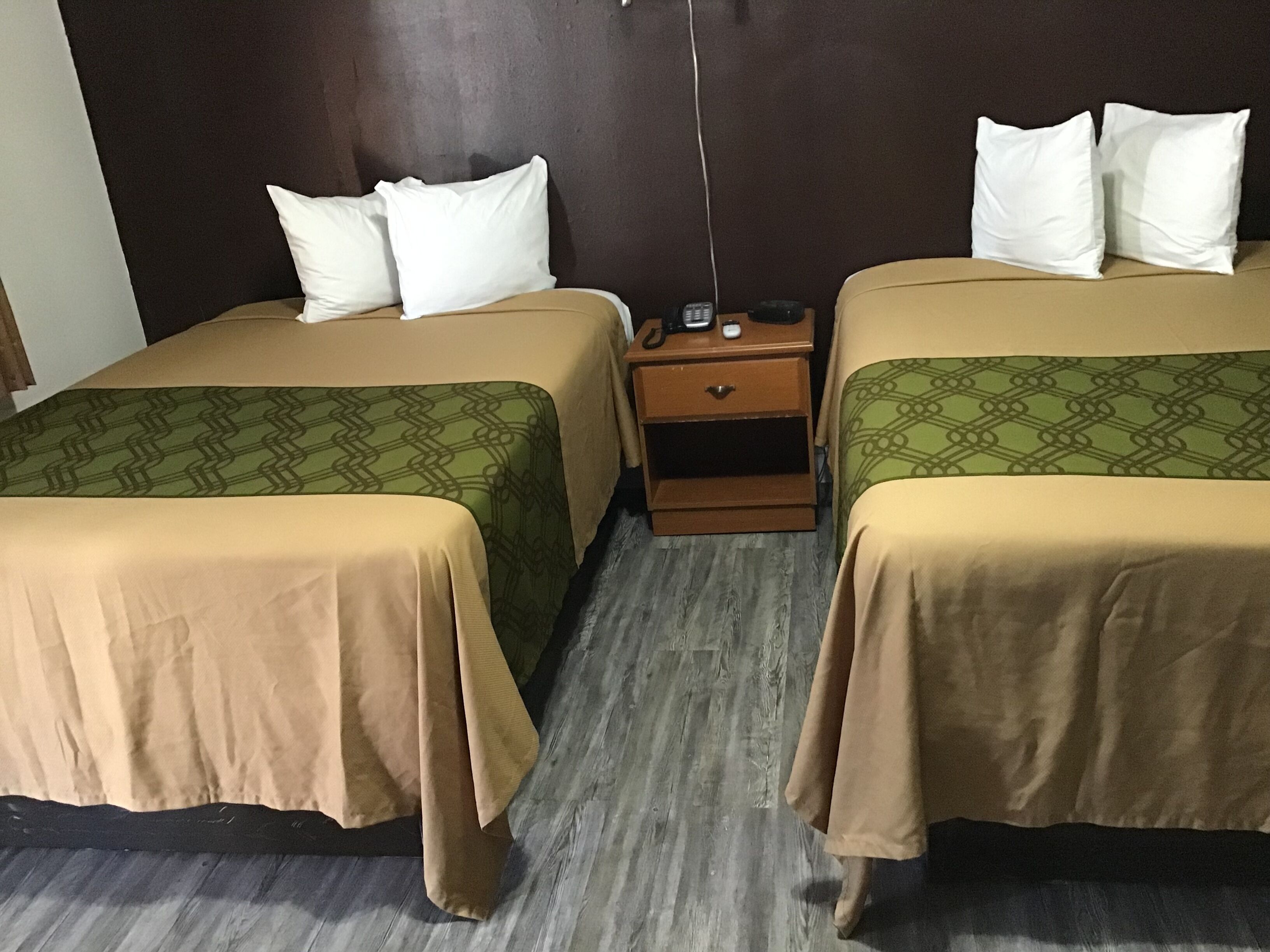 Quarto de hotel com duas camas, cada uma com colcha verde estampada e almofadas brancas. Há uma mesa de cabeceira de madeira entre as camas, com um telefone e uma pequena mesa. O quarto tem uma cabeceira escura e piso de madeira.