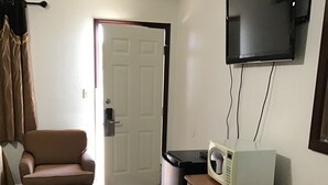 Habitación pequeña con sofá marrón, microondas sobre una mesa de madera, televisor montado en la pared y una puerta que da a otra habitación.