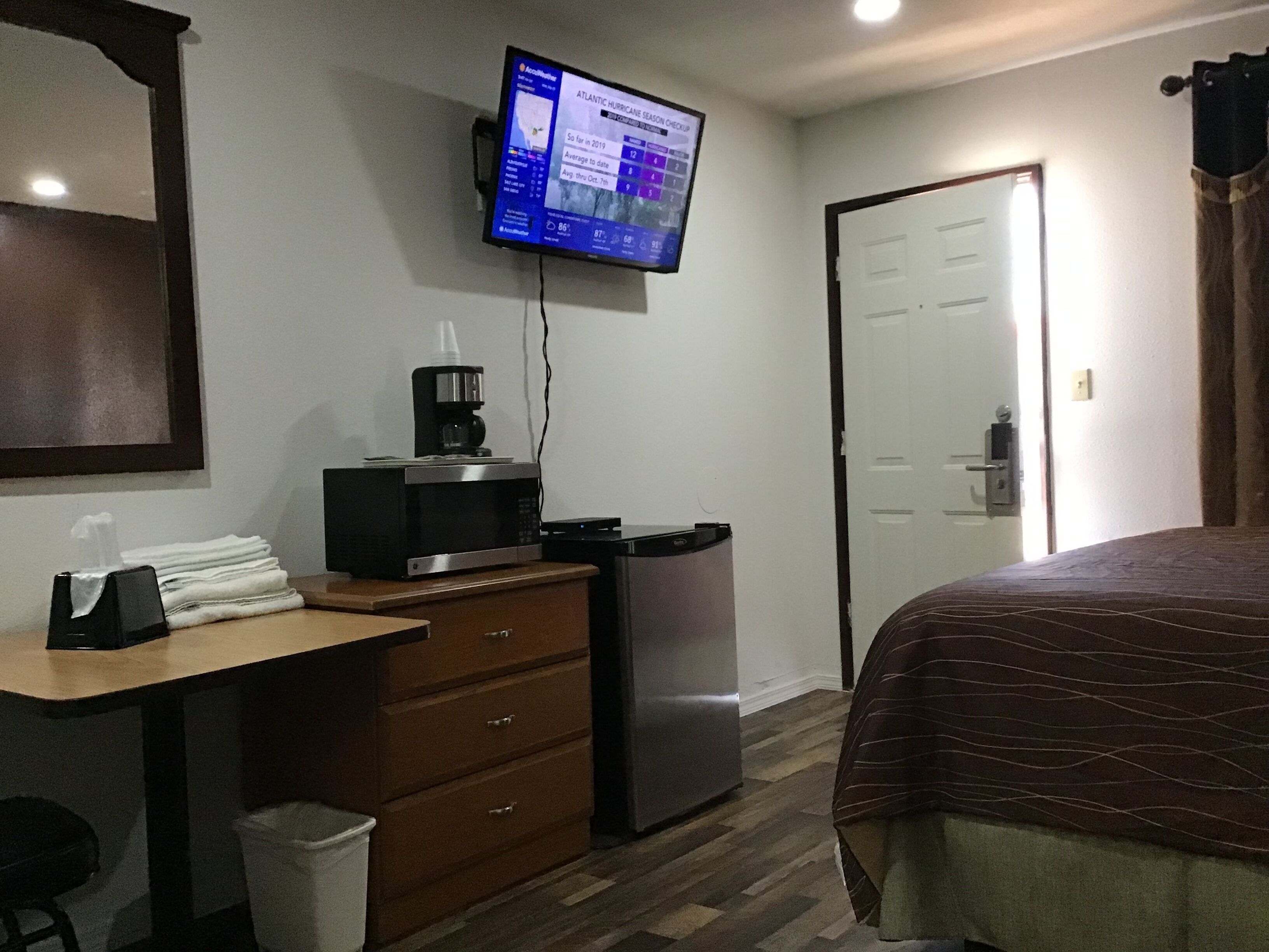 A hotel room with a bed, a desk with a computer monitor displaying hurricane information, a coffee maker, a refrigerator, and a trash bin.