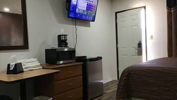 A hotel room with a bed, a desk with a computer monitor displaying hurricane information, a coffee maker, a refrigerator, and a trash bin.
