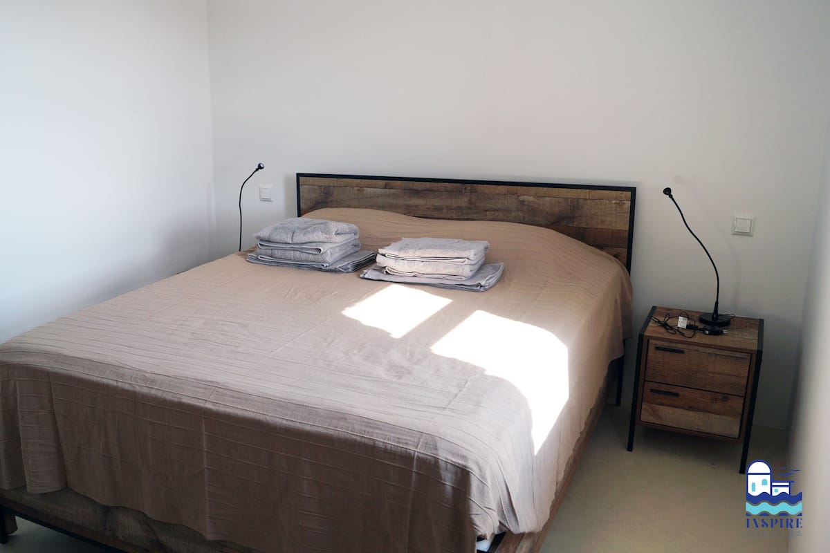 Ein Einzelbett mit Holz-Kopfstück, ein Nachttisch mit Lampe sowie eine Wandsteckdose.