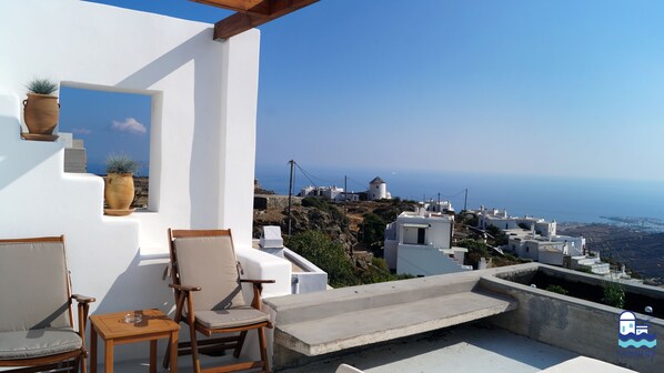 A terrace with two wooden chairs and a small table, overlooking a coastal village with white buildings and a clear blue sky.