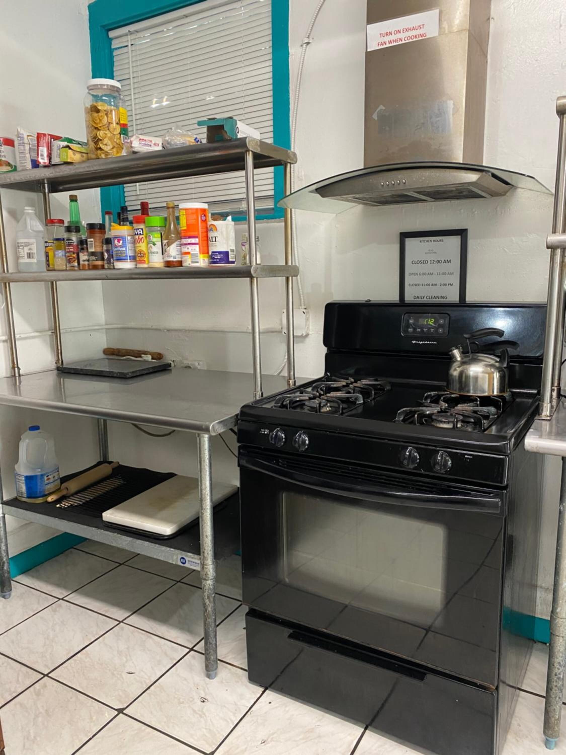 Una cucina con piano cottura a gas, scaffali in acciaio inossidabile e diversi alimenti, e un cartello che indica gli orari di apertura della cucina.