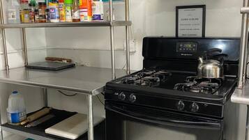 Una cucina con piano cottura a gas, scaffali in acciaio inossidabile e diversi alimenti, e un cartello che indica gli orari di apertura della cucina.