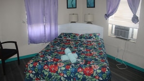A bedroom with a floral bedspread, two bedside lamps, a window with an air conditioner, and purple curtains. - Hoosville Hostel (Florida City)