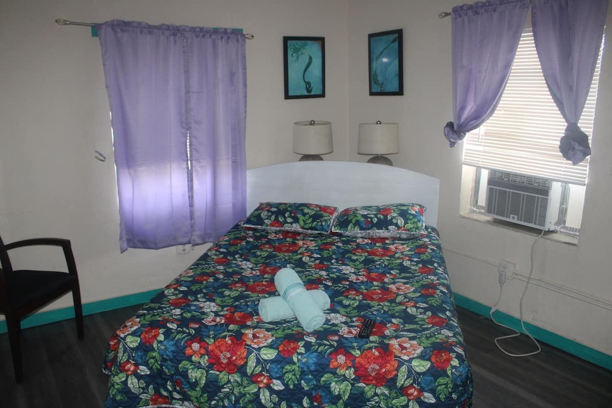 A bedroom with a floral bedspread, two bedside lamps, a window with an air conditioner, and purple curtains.