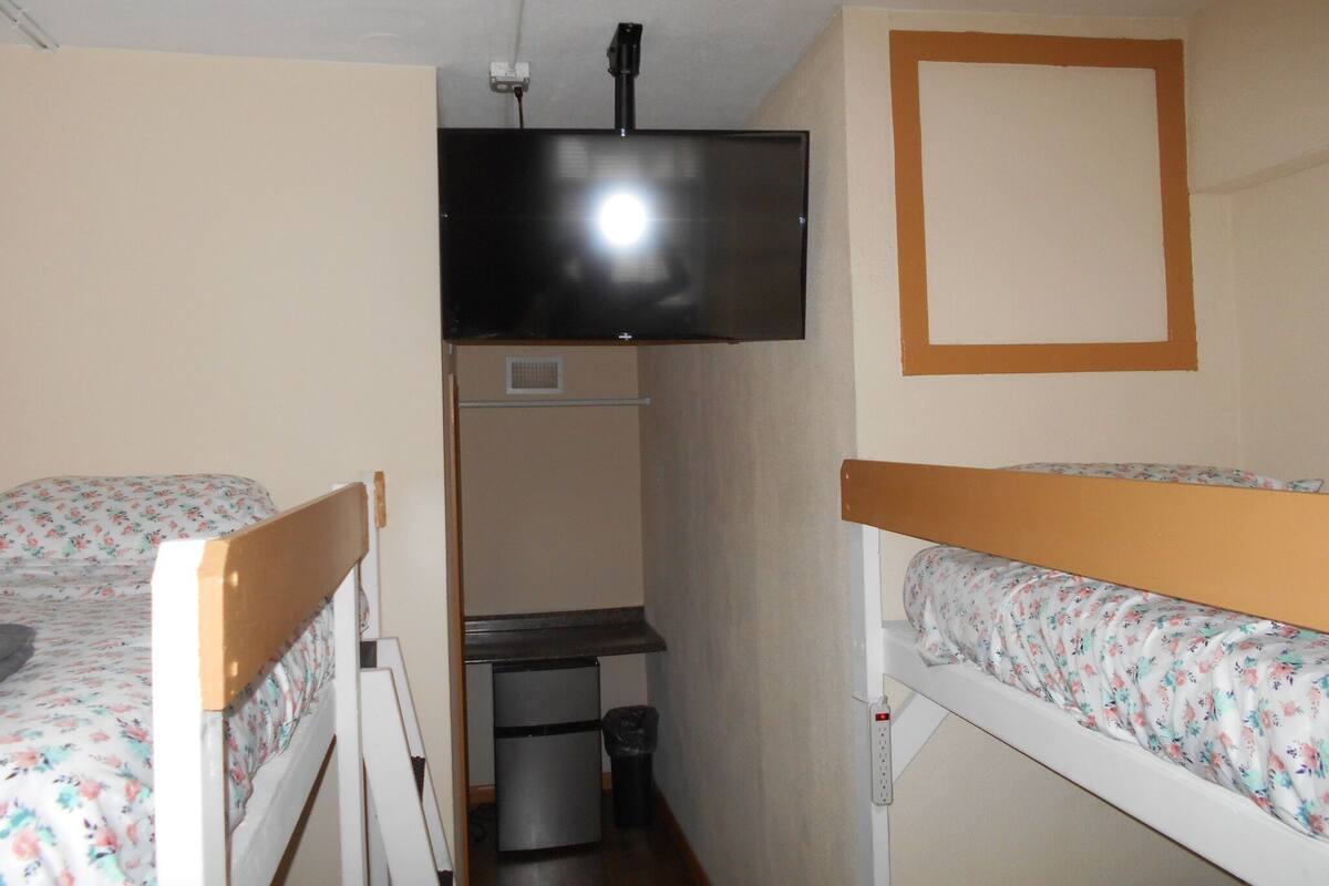 A small, compact room with bunk beds, a television mounted on the wall, a small kitchenette area, and a ceiling-mounted light fixture.