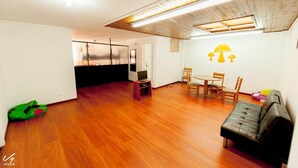 Un salón espacioso con suelo de madera, un sofá de cuero negro, una mesa de comedor con sillas y un televisor sobre un mueble.