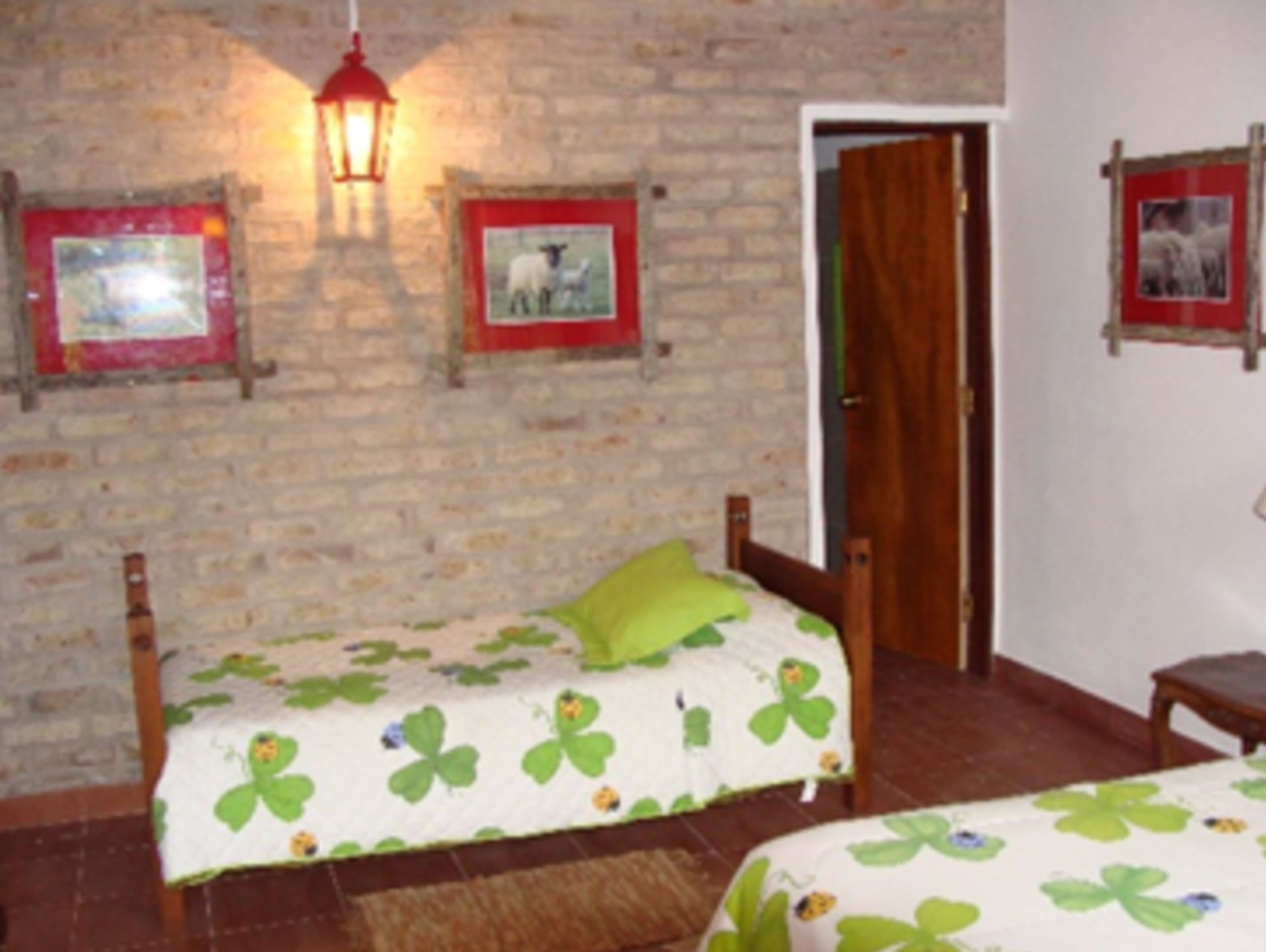 Una stanza con due letti di legno, ognuno con una coperta verde e bianca decorata con motivi di trifoglio. Si vede un quadro con cornice rossa appeso al muro, una lanterna penzolante e una porta di legno.