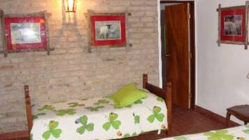 Una stanza con due letti di legno, ognuno con una coperta verde e bianca decorata con motivi di trifoglio. Si vede un quadro con cornice rossa appeso al muro, una lanterna penzolante e una porta di legno.