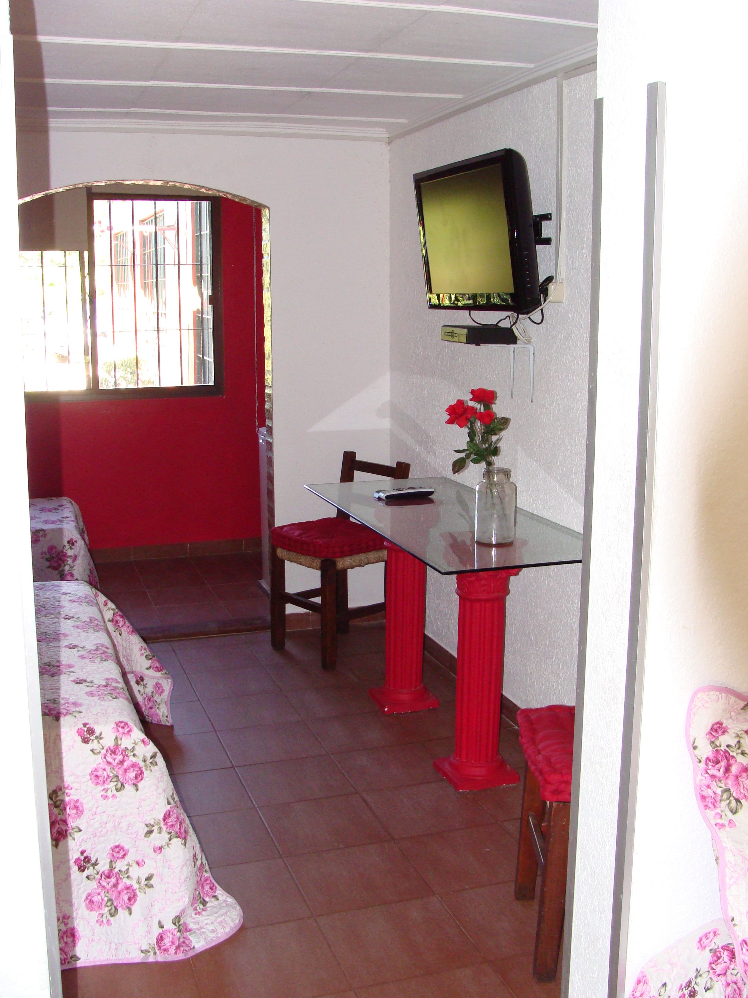 Una habitación pequeña con una mesa de comedor, un televisor en la pared y una ventana con rejas.